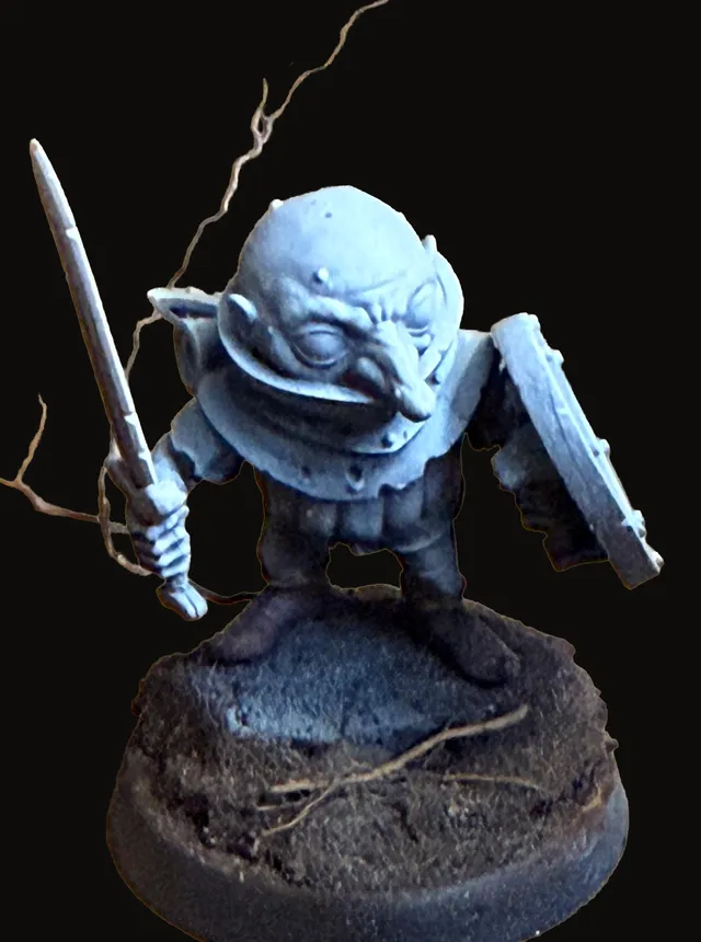 An unpainted miniature of Rollo — a small svirfneblin with a wide bald head and oversized ears, sword raised in one hand and a round shield held at the ready in the other, standing in a battle-ready crouch on a dark textured round base