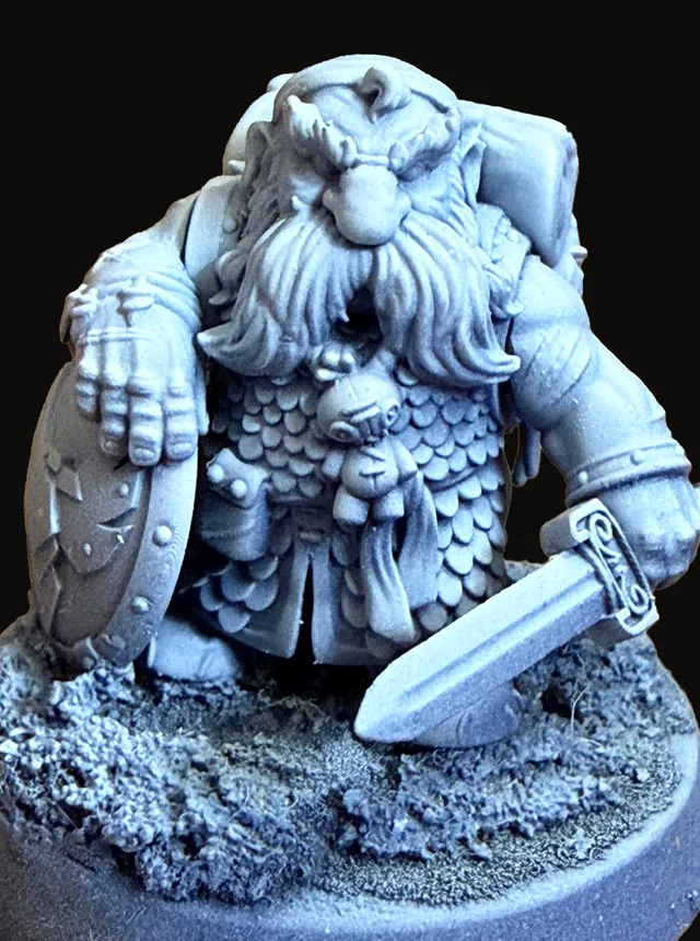 An unpainted resin miniature of Thorek — a stocky, bearded dwarven figure in heavy scaled armor and a heavy fur cloak, holding a sword at rest across his body, standing on an oval scenic base