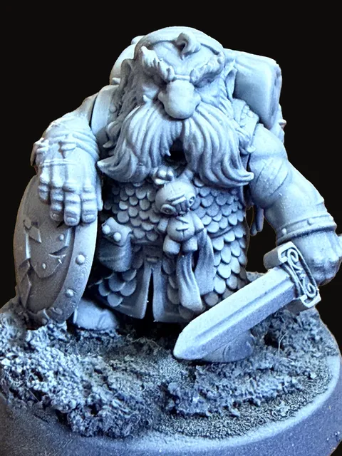 An unpainted resin miniature of Thorek — a stocky, bearded dwarven figure in heavy scaled armor and a heavy fur cloak, holding a sword at rest across his body, standing on an oval scenic base